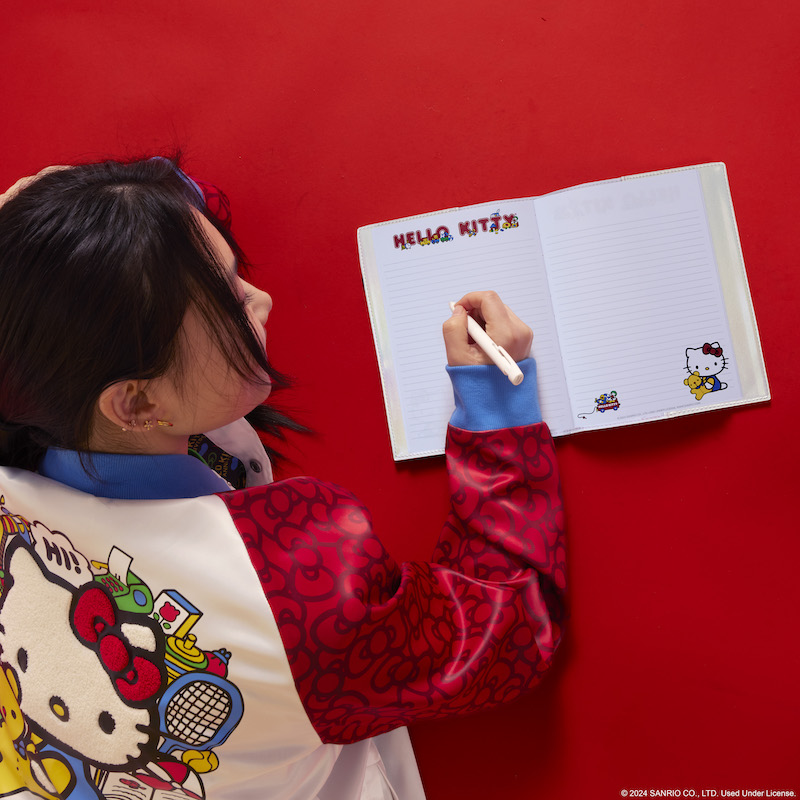 Woman wearing the Loungefly Hello Kitty Souvenir Jacket and laying face down on a red floor and journaling in the Hello Kitty 50th Anniversary Cosplay Pearlescent Refillable Stationery Journal 